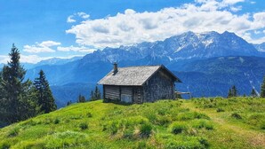 Una pequeña cabaña de madera en una colina cubierta de hierba, con montañas al fondo.