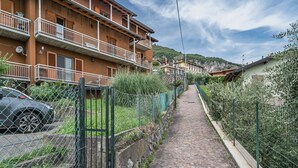 Un étroit sentier pavé, bordé de bâtiments résidentiels dotés de balcons, avec une voiture garée sur la gauche et une clôture verte sur la droite.