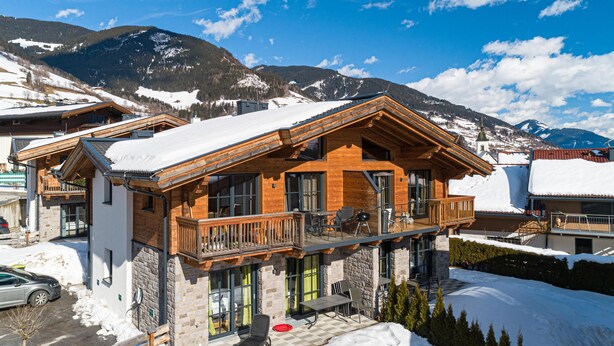 A wooden chalet with a balcony, stone walls, and a view of snow-covered mountains.
