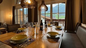 A dining area with a wooden table set for a meal, large windows with a mountain view, and hanging pendant lights.