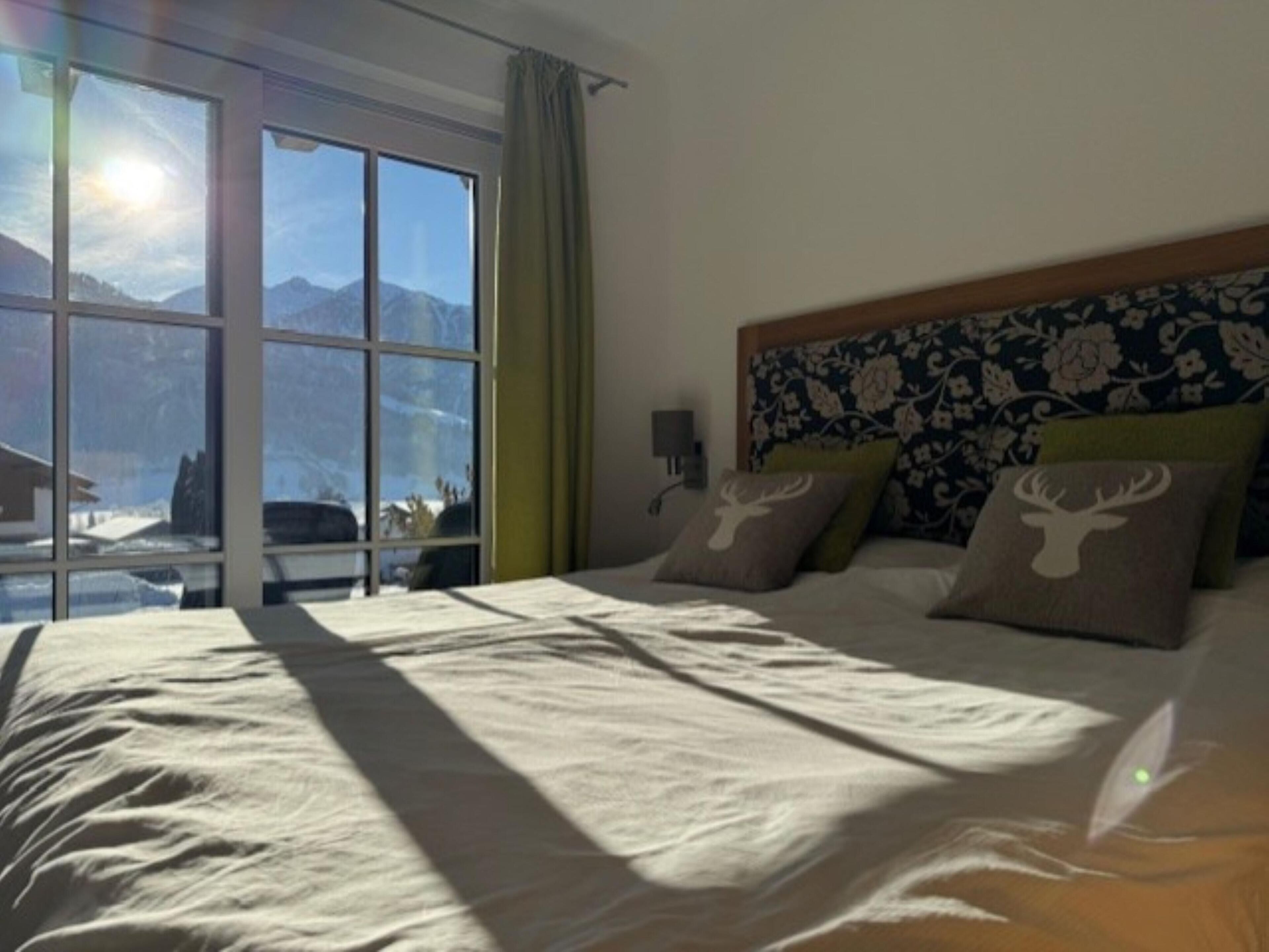 A hotel room with a large bed, a headboard with floral patterns, and a view of mountains through the window.