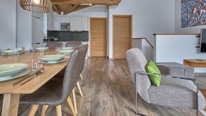 A modern living space with a wooden dining table, chairs, a sofa, and a kitchen area in the background.