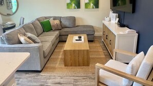 A modern living room with a grey sectional sofa, a wooden coffee table, and a white armchair. There are three green leaf paintings on the wall, a flat-screen TV, and a small cabinet with a phone. - Your Perfect Family Retreat! (Kissimmee)