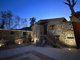 Un edificio de piedra con una escalera que conduce a una puerta, asientos exteriores con sillas y una vista de árboles al fondo.