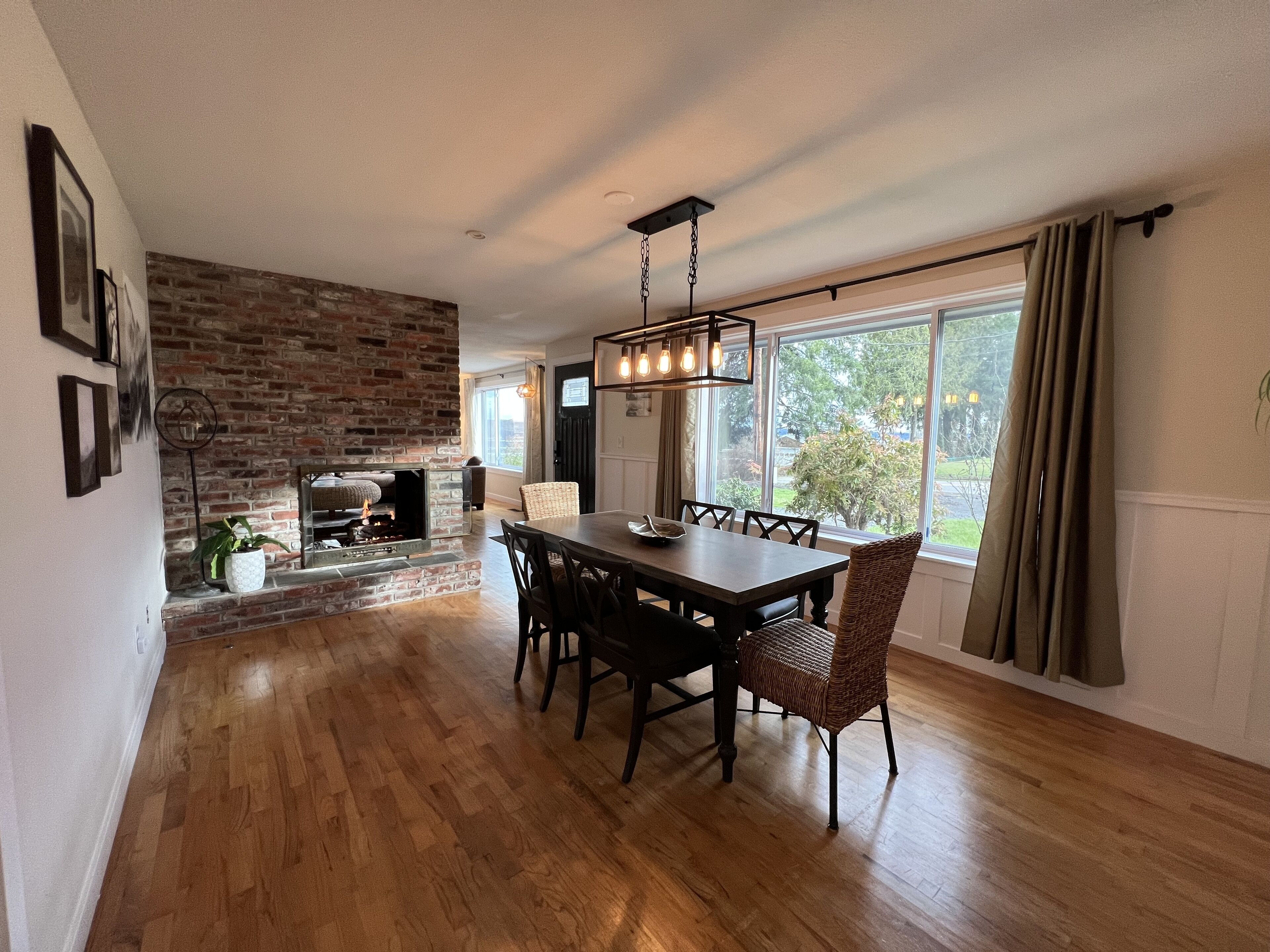 Un coin repas agrémenté d’une cheminée en briques, de parquet et d’une grande fenêtre avec des rideaux.