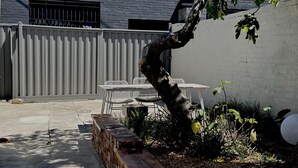 A patio area with a white table and chairs, a brick planter, and a tree.
