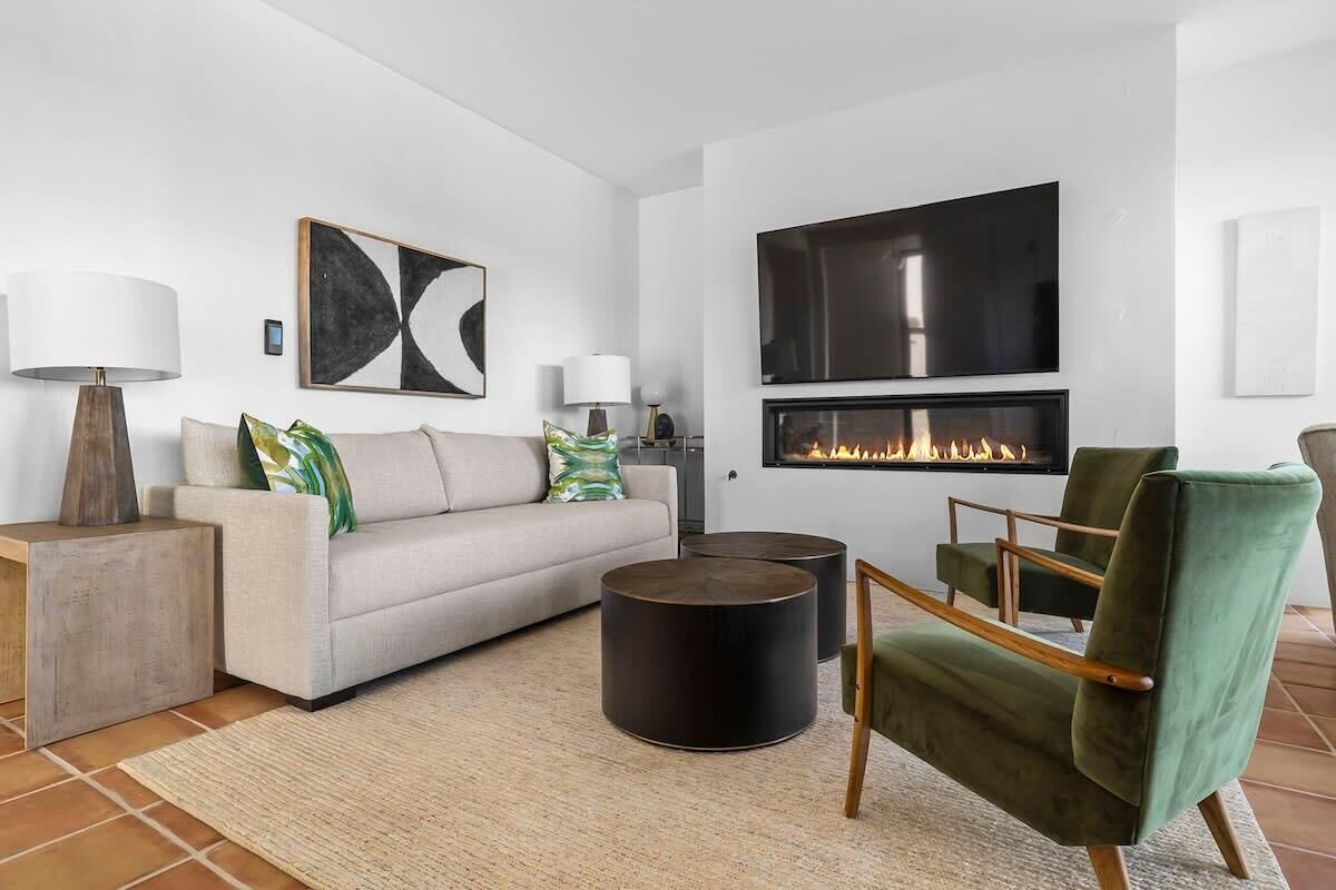 A modern living room with a sofa, two side tables, a green armchair, a fireplace, and a large TV mounted on the wall.