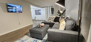 Un salon moderne avec un canapé gris, un pouf gris et un téléviseur  à écran plat fixé au mur.
