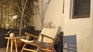 Una terraza de madera con una silla de mimbre y una pequeña mesa, una planta en maceta y bambú al fondo.
