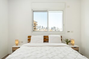 Ein Schlafzimmer mit Blick auf die Stadt, einem Holzkopfende, zwei Nachttischlampen und einer weißen Bettdecke.