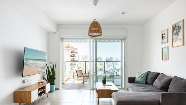 A modern living room with a grey sofa, a wooden TV stand, and a large window offering a city view. - MAMAD - Sea Breeze Netanya Retreat by Sea N Rent (Netanya)