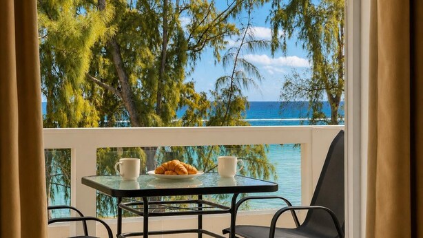 Un balcone con tavolo, sedie e un cornetto, affacciato su una spiaggia con alberi e l'oceano.