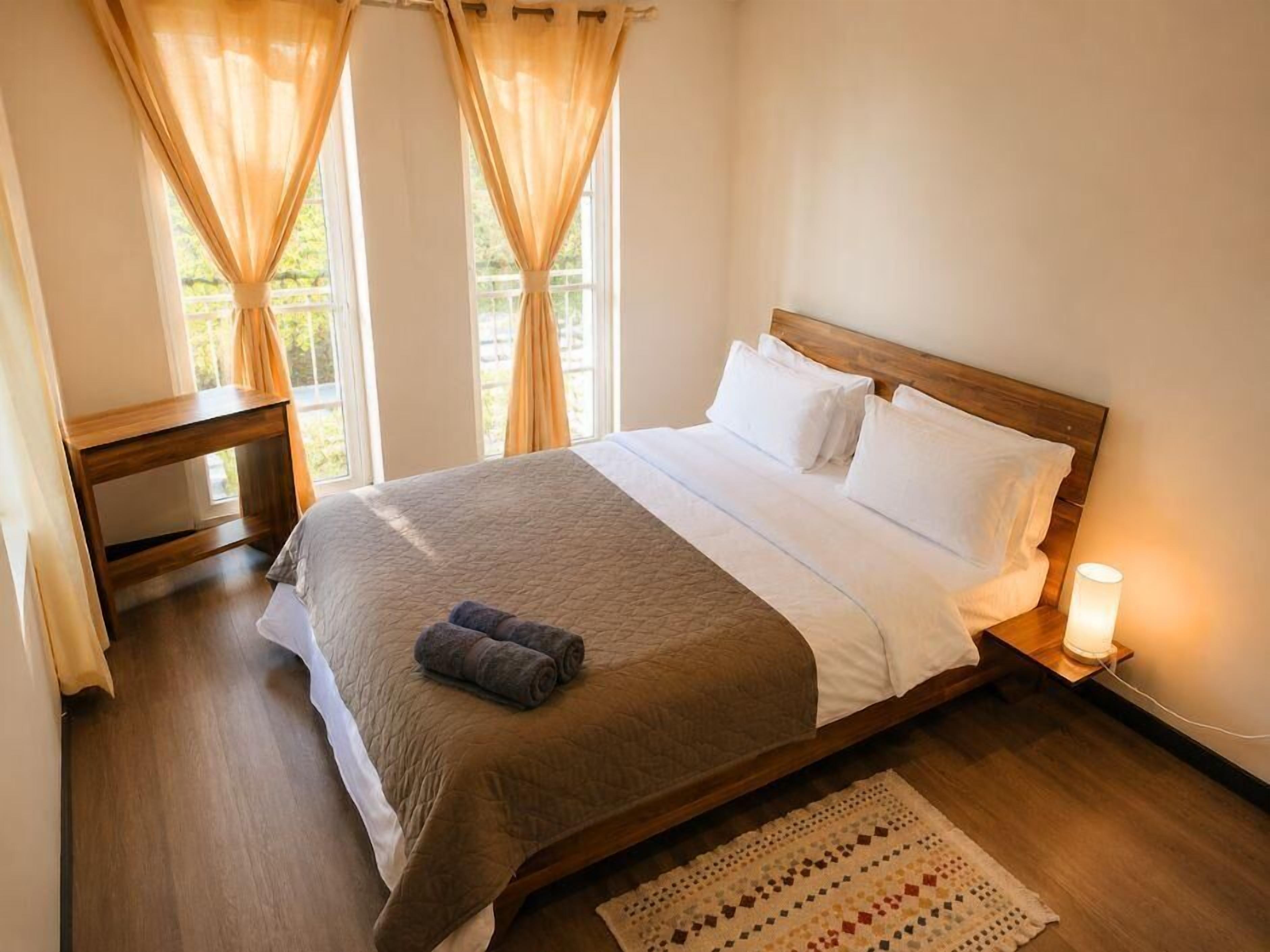 A neatly made bed with a wooden headboard, a bedside table with a lamp, and a window with curtains.