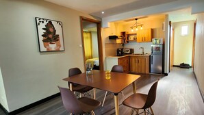 Una zona pranzo con tavolo in legno e quattro sedie, una cucina con mobili in legno ed elettrodomestici moderni, e una pianta in vaso appesa al muro.
