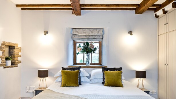 A bedroom with a large bed, two bedside lamps, a window with a view, and a stone wall feature. - The Crewe & Harpur pub, a unique exclusive self catering venue, sleeps up to 34. (Buxton)