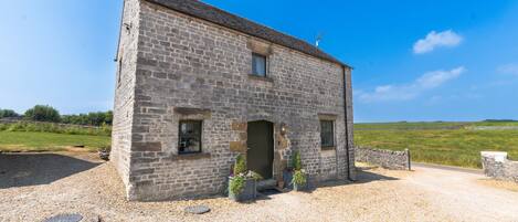 A stone building with a gravel driveway, a small garden area with potted plants, and a clear blue sky.