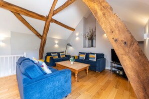 A loft-style living room with exposed wooden beams, a blue sofa set, a wooden coffee table, and a flat-screen TV.