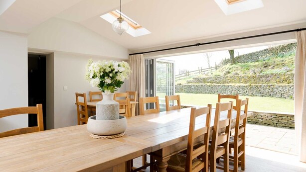 A dining area with a wooden table and chairs, a vase with flowers, and a view of a stone wall and greenery outside. - Lapwing Barns, large rural farmhouse with games room, perfect for all types of groups. (Longnor)