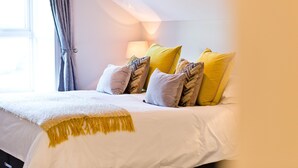 Una cama bien hecha con una colcha blanca y amarilla, varios almohadones con distintos diseños y una lámpara en una mesita de noche.