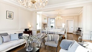 Amplia sala de estar con comedor, con candelabro, paredes blancas y un ventanal grande.