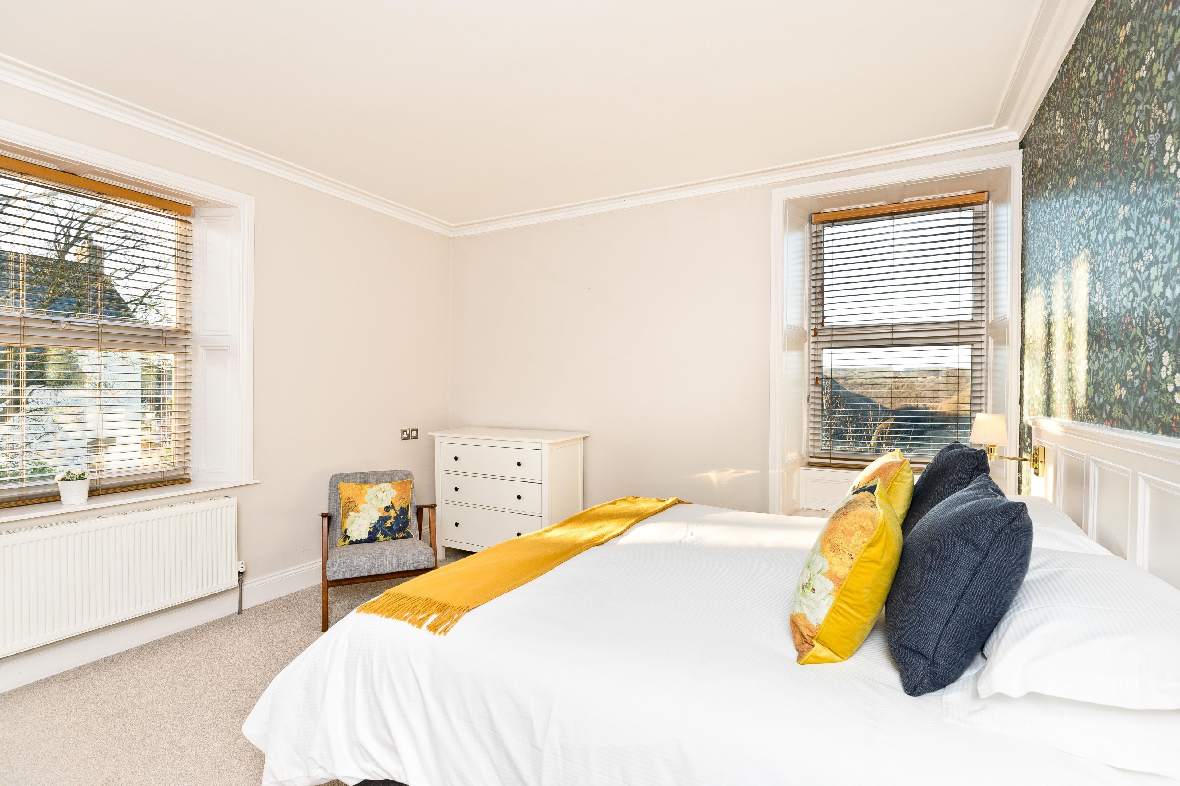 A bedroom with a large bed, a nightstand, a chair, and two windows with blinds.