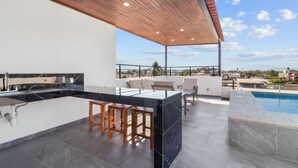 Une cuisine extérieure moderne avec piscine, comprenant un bar avec des tabourets, un espace détente avec une table et des chaises, et une vue sur le paysage environnant.
