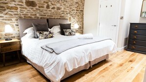 Une chambre avec un mur en pierre, un lit avec une literie blanche, une table de chevet en bois et une commode avec des tiroirs.