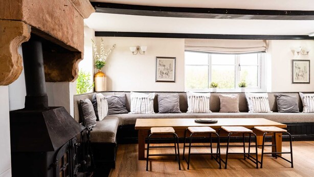 A cozy living room with a built-in fireplace, a wooden dining table with four chairs, a large window with a view, and a sofa with patterned cushions.