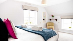 A bedroom with a bed, pillows, a blue blanket, a chair, a mirror, and two windows with blinds.
