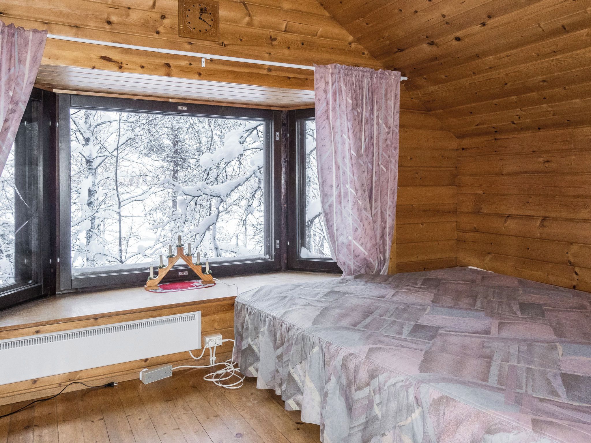 Mökkityyppinen huone, jossa on sänky, ikkuna verhoilla, patteri ja puinen katto.