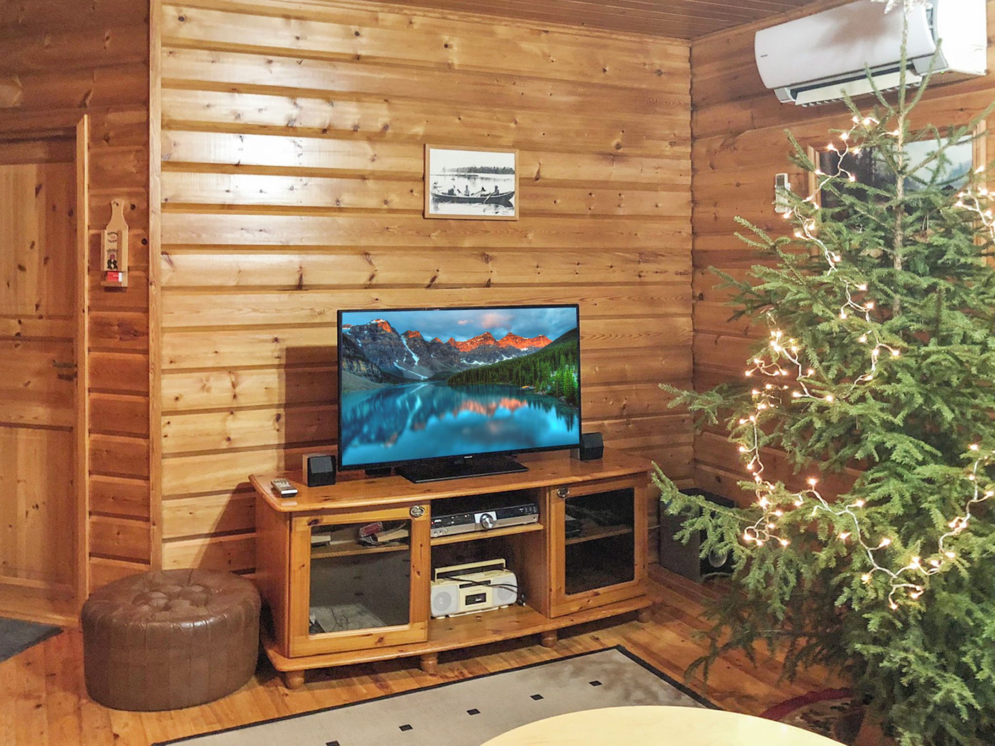 Un intérieur en bois avec un téléviseur posé sur un meuble en bois, un petit sapin de Noël illuminé et un tableau encadré accroché au mur.