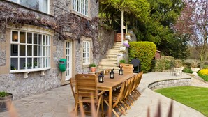 Une terrasse en pierre avec une table et des chaises en bois, entourée de plantes en pot et d’un mur de pierre.