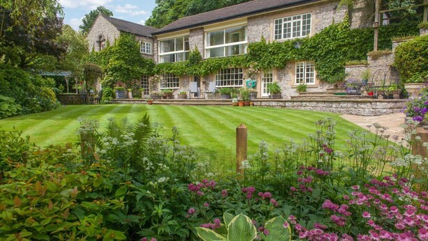 Un bâtiment en pierre, doté d’une pelouse bien entretenue et agrémenté de diverses plantes à fleurs au premier plan.