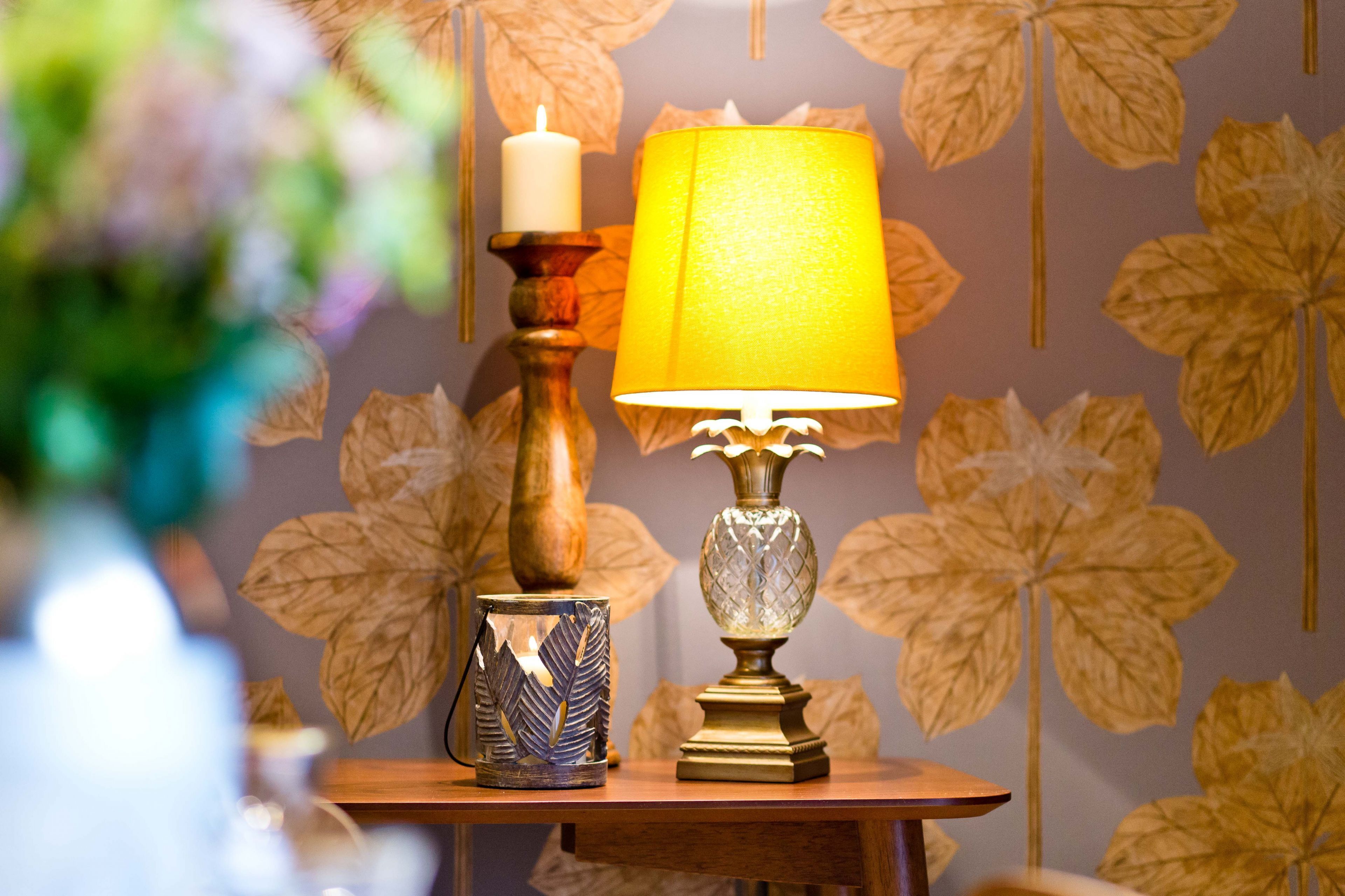 Une table avec une bougie et une lampe, placée devant un mur orné d’un motif de feuilles.