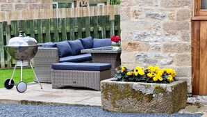A wicker sofa with cushions, a barbecue grill, and a planter with yellow flowers. - Modern 5 bedroom family friendly barn conversion in the heart of Bakewell. (Bakewell)