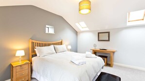 A modern bedroom with a sloped ceiling, a wooden bed, a bedside table with a lamp, a desk, and a mirror. - Modern 5 bedroom family friendly barn conversion in the heart of Bakewell. (Bakewell)