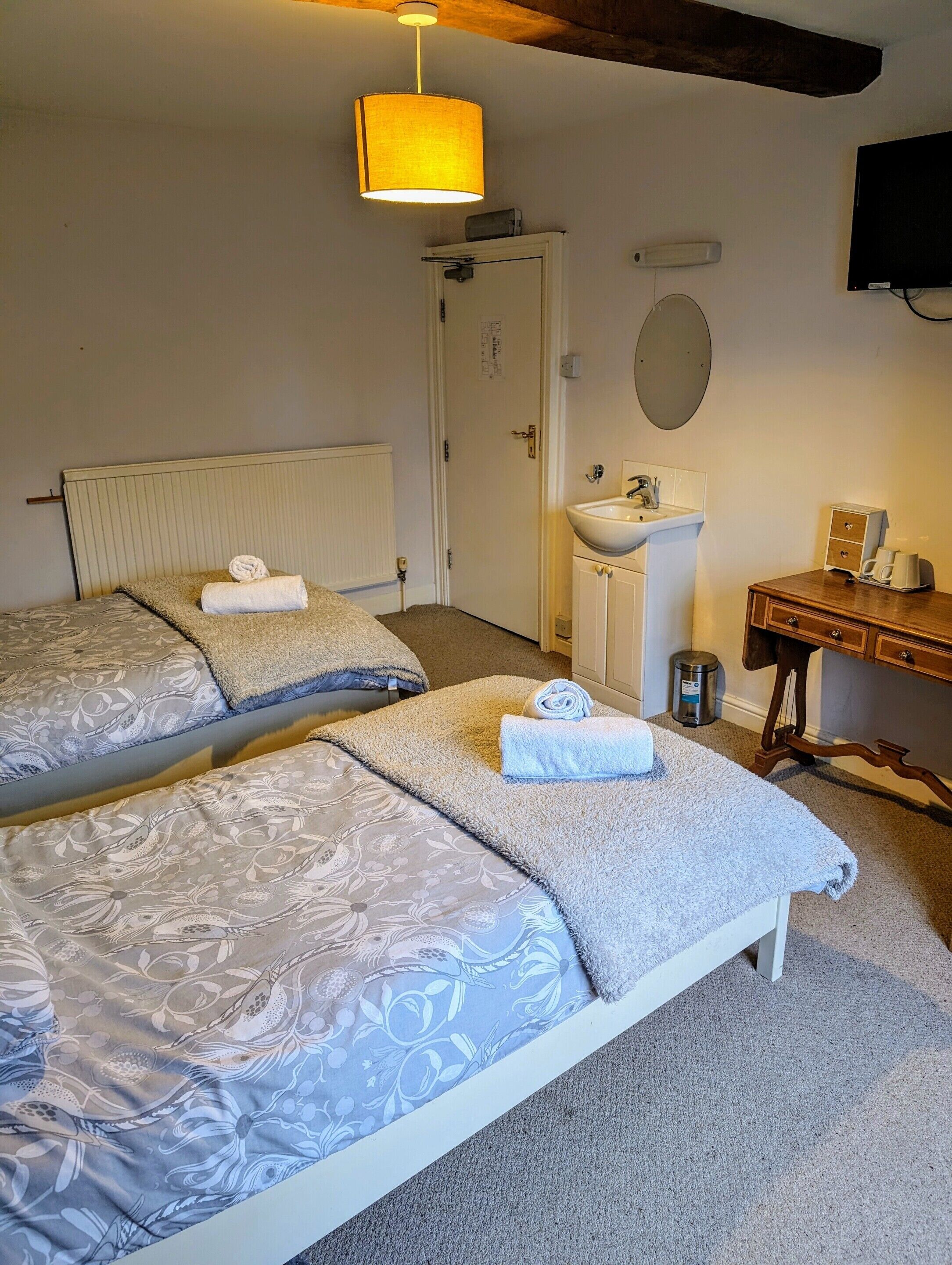 A hotel room with two beds, a bedside table, a lamp, a sink, and a television.