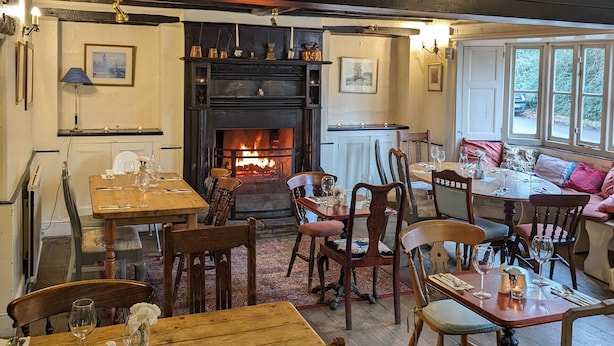 A cozy dining area with wooden tables and chairs, a fireplace, and a view through large windows. - Bartholomew Arms (Towcester)