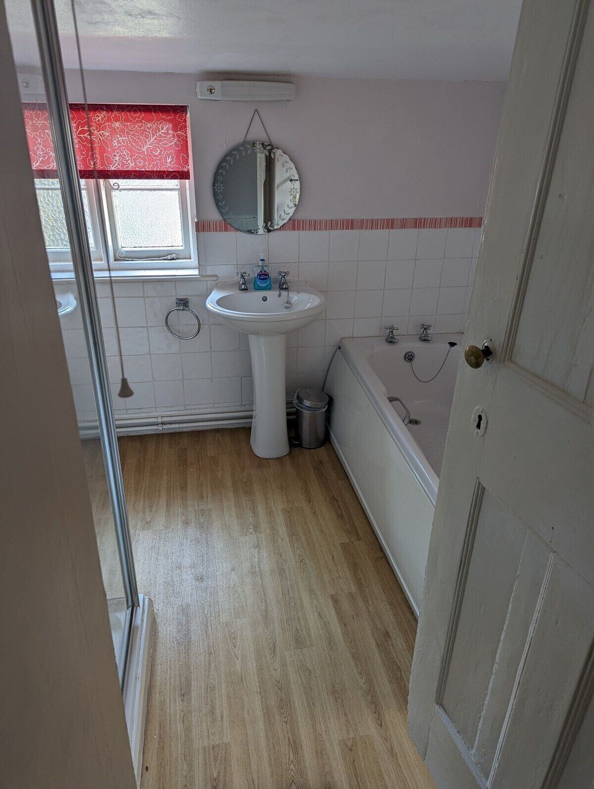 A bathroom with a white sink, a mirror, a red window valance, a white bathtub, and a wooden floor.