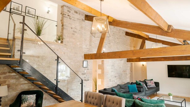A spacious loft with exposed wooden beams, a staircase, a living area with a sofa and cushions, and a dining table. - Large luxury farmhouse retreat situated in a rural hamlet, perfect for large gatherings of up to 50 (Matlock)