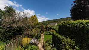 A stone pathway leading through a lush garden with various plants and a wooden fence on one side. - A cosy country cottage in the village of Curbar with amazing views of Curbar Edge (Curbar)