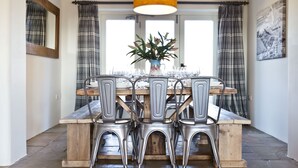 Un coin repas avec une table en bois dressée pour quatre personnes, entourée de chaises en métal, d’un vase rempli de fleurs et de rideaux à carreaux.