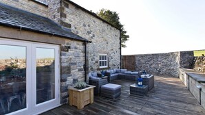 Un bâtiment en pierre doté d’une grande porte vitrée, d’une terrasse en bois et d’un espace extérieur aménagé pour s’asseoir.