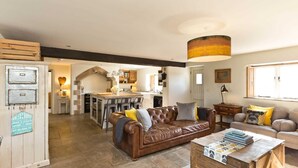 Un salon chaleureux avec un canapé en cuir marron, une table basse avec des livres, et une cuisine en arrière-plan.