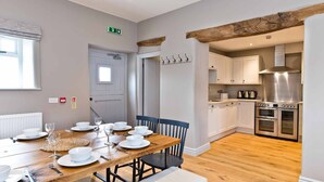 A dining area with a wooden table set for six, white chairs, and a window with blinds. - Stylish Farmhouse with stunning views over rolling hills and fabulous games room. (Belper)
