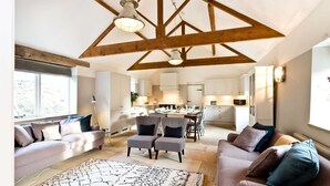 A spacious living area with a high ceiling, exposed wooden beams, a dining area with a table and chairs, and a kitchen in the background. - Collection of 4 charming barns offering group accommodation for 16, close to Bakewell (Bakewell)