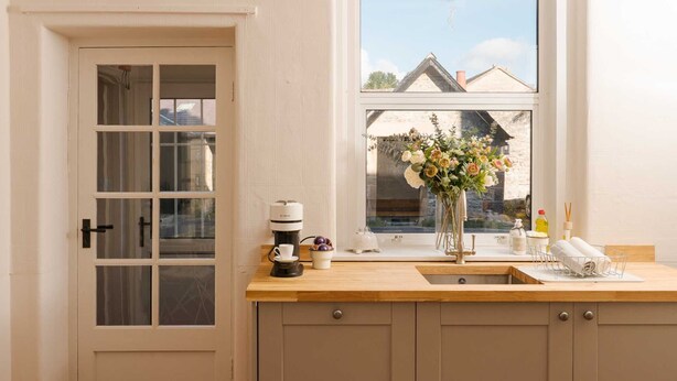 A kitchen with a wooden countertop, a coffee maker, a vase with flowers, and a view of buildings through the window. - Quaint 2 bedroom cottage in the picturesque village of Ashford-in-the-Water. (Bakewell)