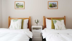 Een hotelkamer met twee bedden, elk met een houten hoofdbord, witte beddengoed en kussens met een bladmotief. Tussen de bedden staat een klein nachtkastje met een bloevas.