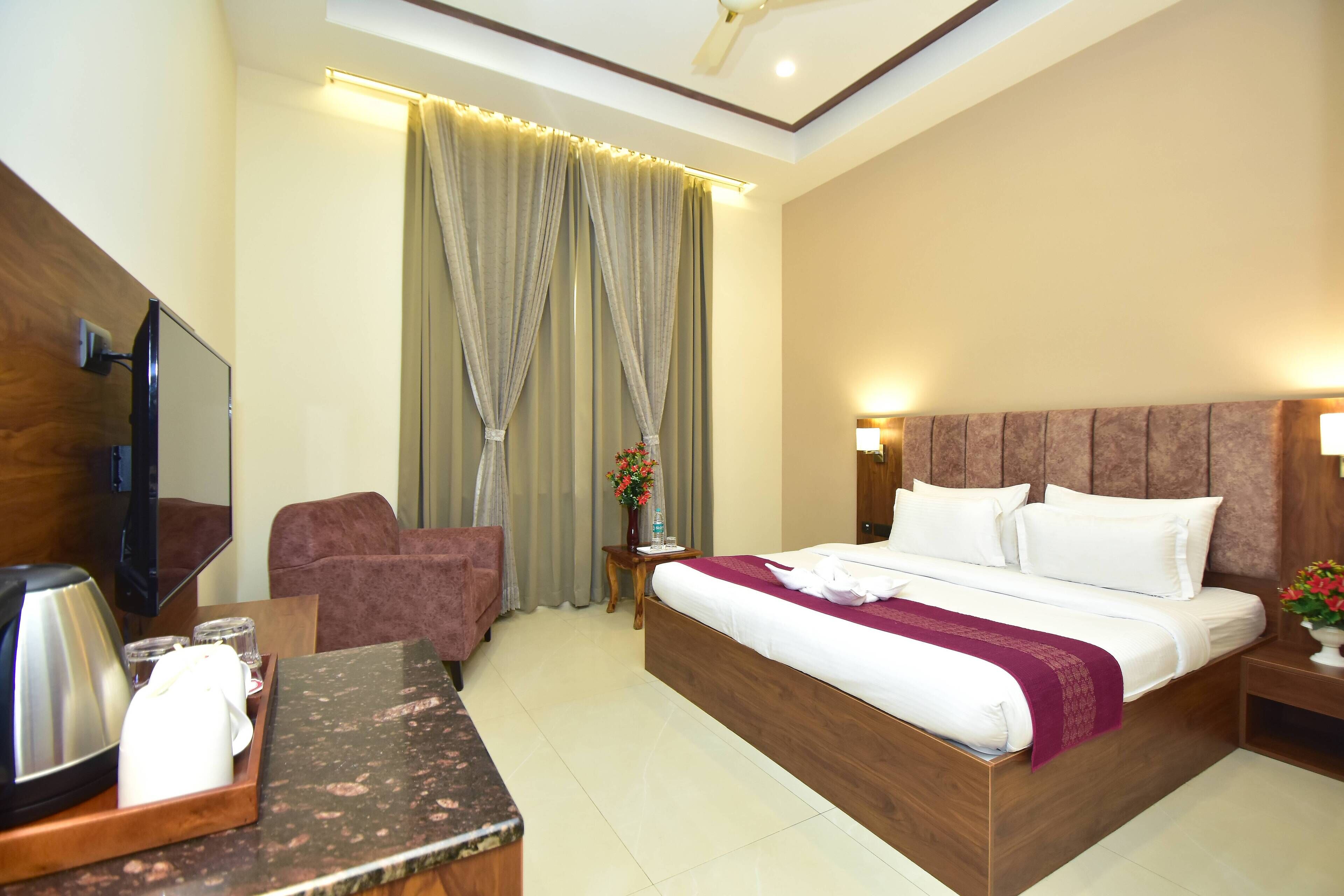 A hotel room with a bed, a TV, a desk with a coffee maker, a chair, and a nightstand with a lamp.
