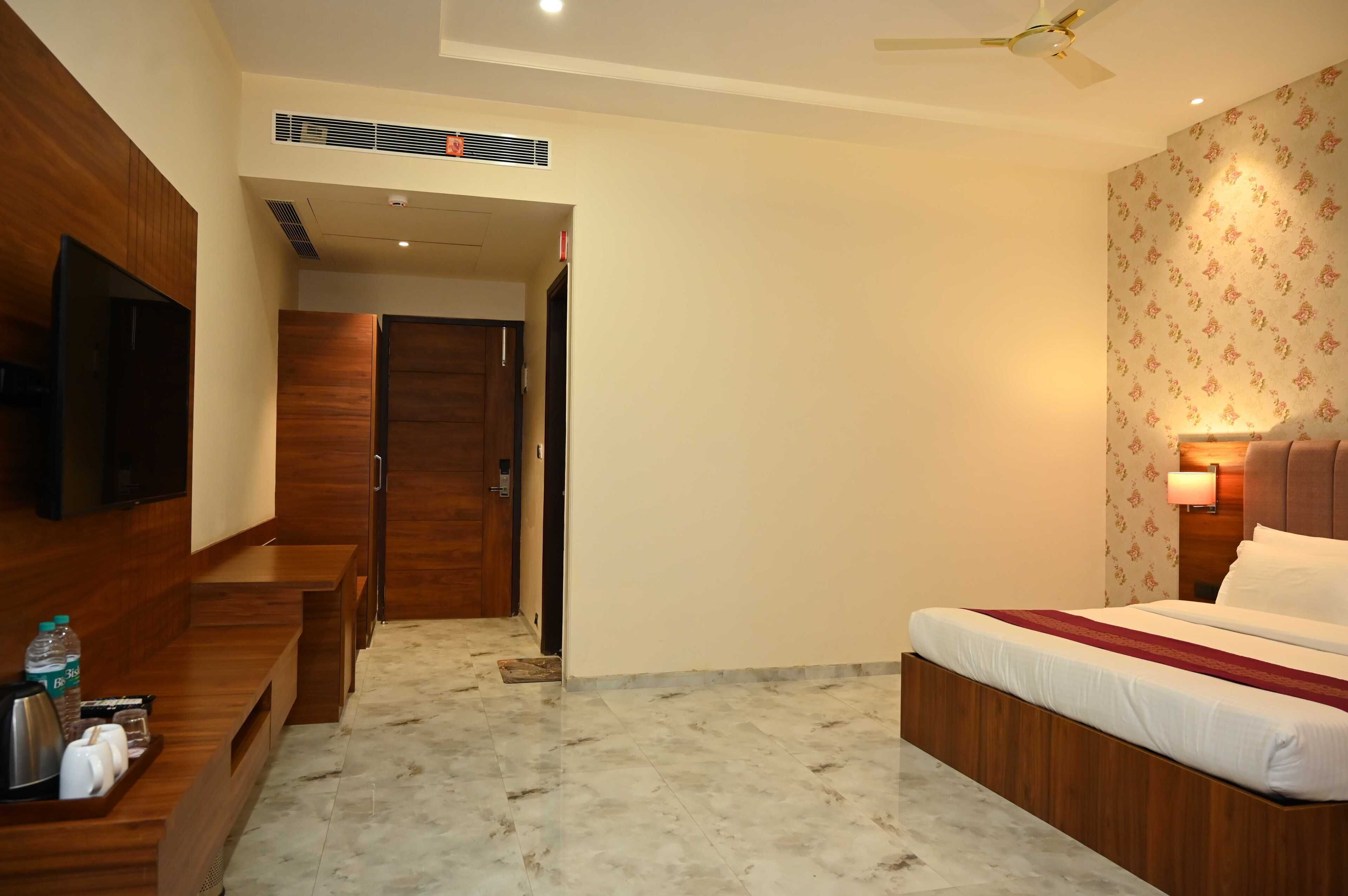 A hotel room with a bed, a TV, a wooden bench, a desk with a water bottle, and a patterned wall.